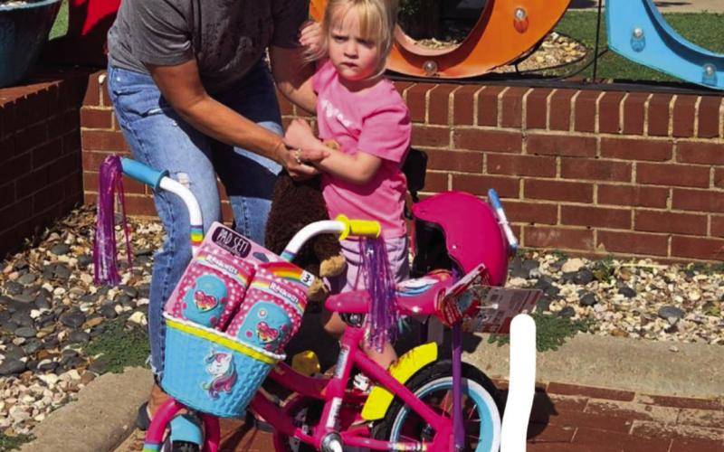 DRAWING WINNER Kennedy Peterson is presented with a 12”girls’ bike, knee and elbow pads, and helmet by Chris Atkinson from the Post Community Recovery Center after the bike drawing on July 4. | DAWN GRAVES PHOTO