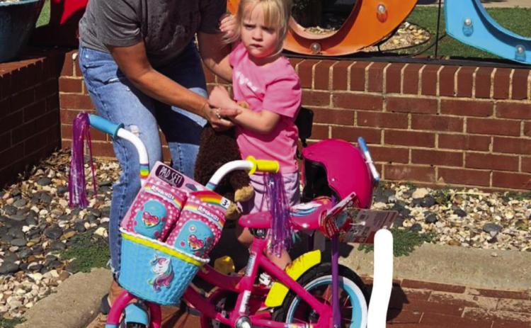 DRAWING WINNER Kennedy Peterson is presented with a 12”girls’ bike, knee and elbow pads, and helmet by Chris Atkinson from the Post Community Recovery Center after the bike drawing on July 4. | DAWN GRAVES PHOTO
