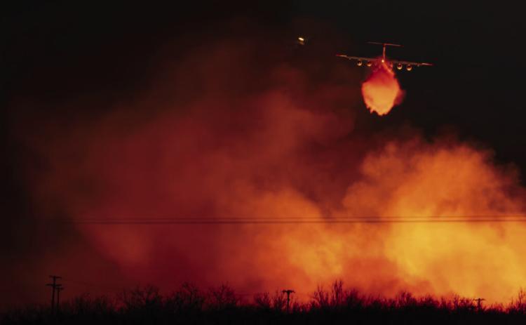 A Texas A&amp;M Forest Service plane drops water on a wall of flames that reaches into the sky as the Smokehouse Creek fire reignited o March 3 near Miami. | TEXAS A&amp;M AGRILIFE SAM CRAFT.